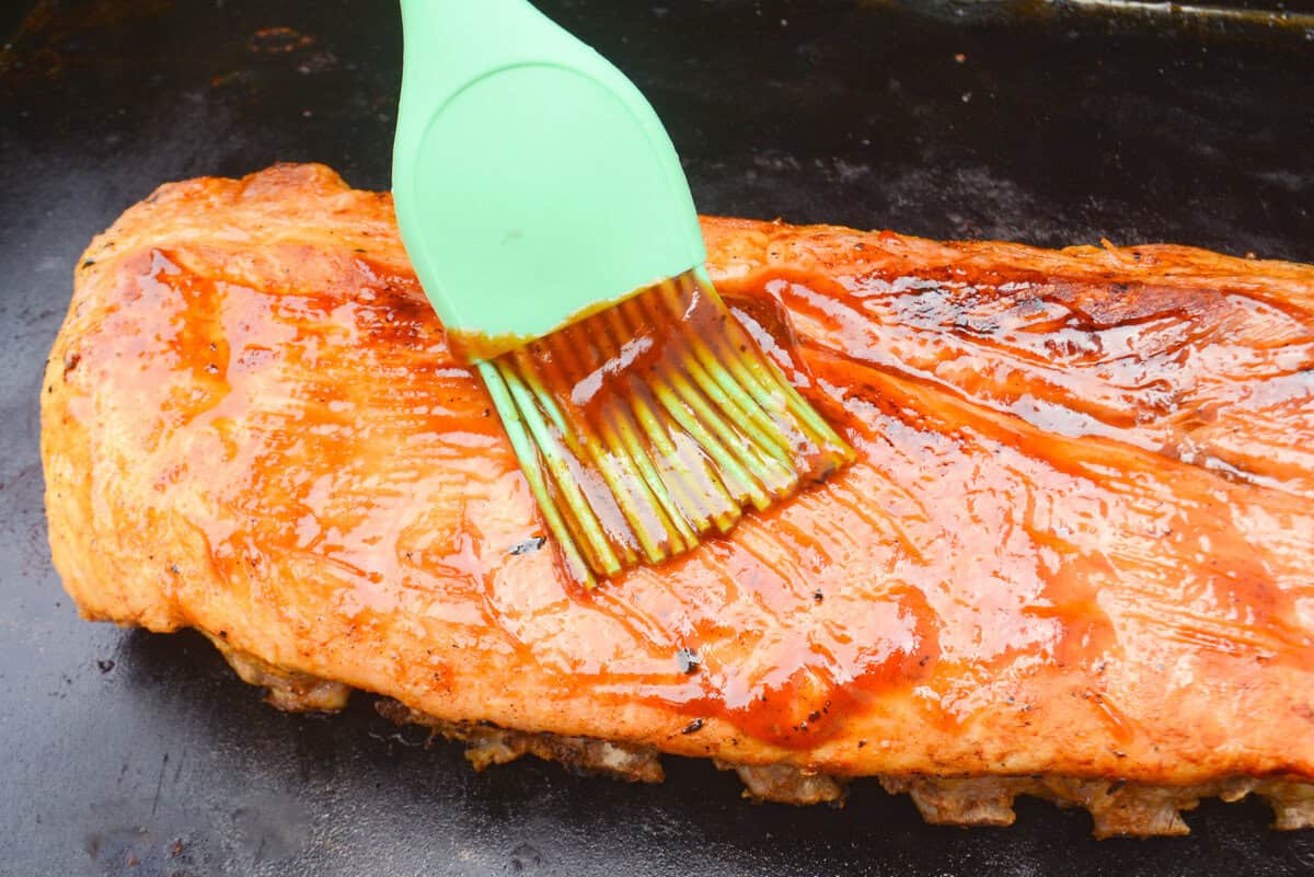 A close up of a green silicone brush adding barbecue sauce to the cooked ribs on the Blackstone