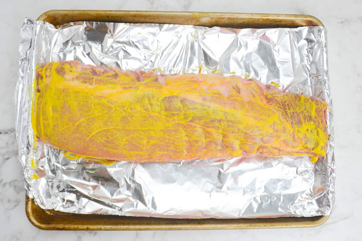 A large baking sheet lined with tinfoil. The uncooked ribs are coated in bright yellow mustard 
