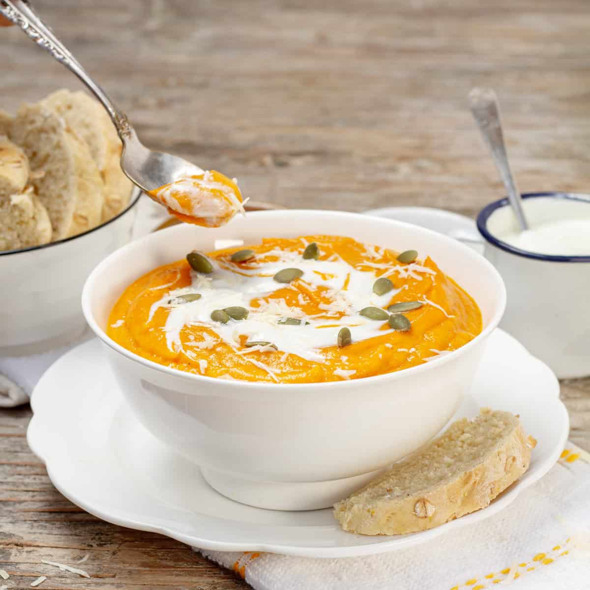 white bowl of roasted carrot soup with a swirl of yogurt and pumpkin seeds, a chunk of bread on the saucer and a jar of yogurt behind