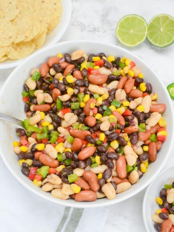 A large white serving bowl bowl filled with Mexican three bean salad. To the side is a bowl of tortilla chips, a halved lime, three slices of jalapeno and a small bowl of the salad