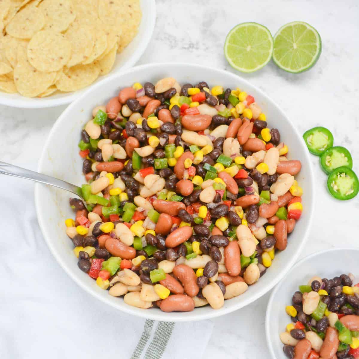 A large white serving bowl bowl filled with Mexican three bean salad. To the side is a bowl of tortilla chips, a halved lime, three slices of jalapeno and a small bowl of the salad