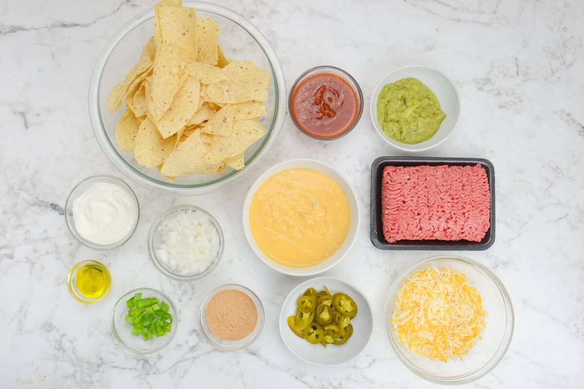 ingredients for making blackstone nachos, including tortilla chips, salsa, guacamole, ground beef, shredded cheese, sliced jalapenos, queso, taco seasoning, diced white onion, diced green onion, olive oil, and sour cream.