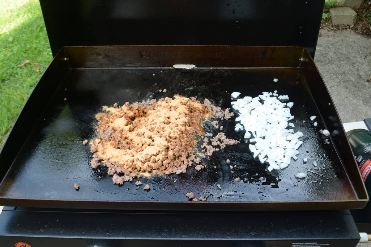 Cooked beef is pushed to the left of the griddle and topped with taco seasoning that has not yet been mixed. Chopped onions sit on the right side of the griddle.