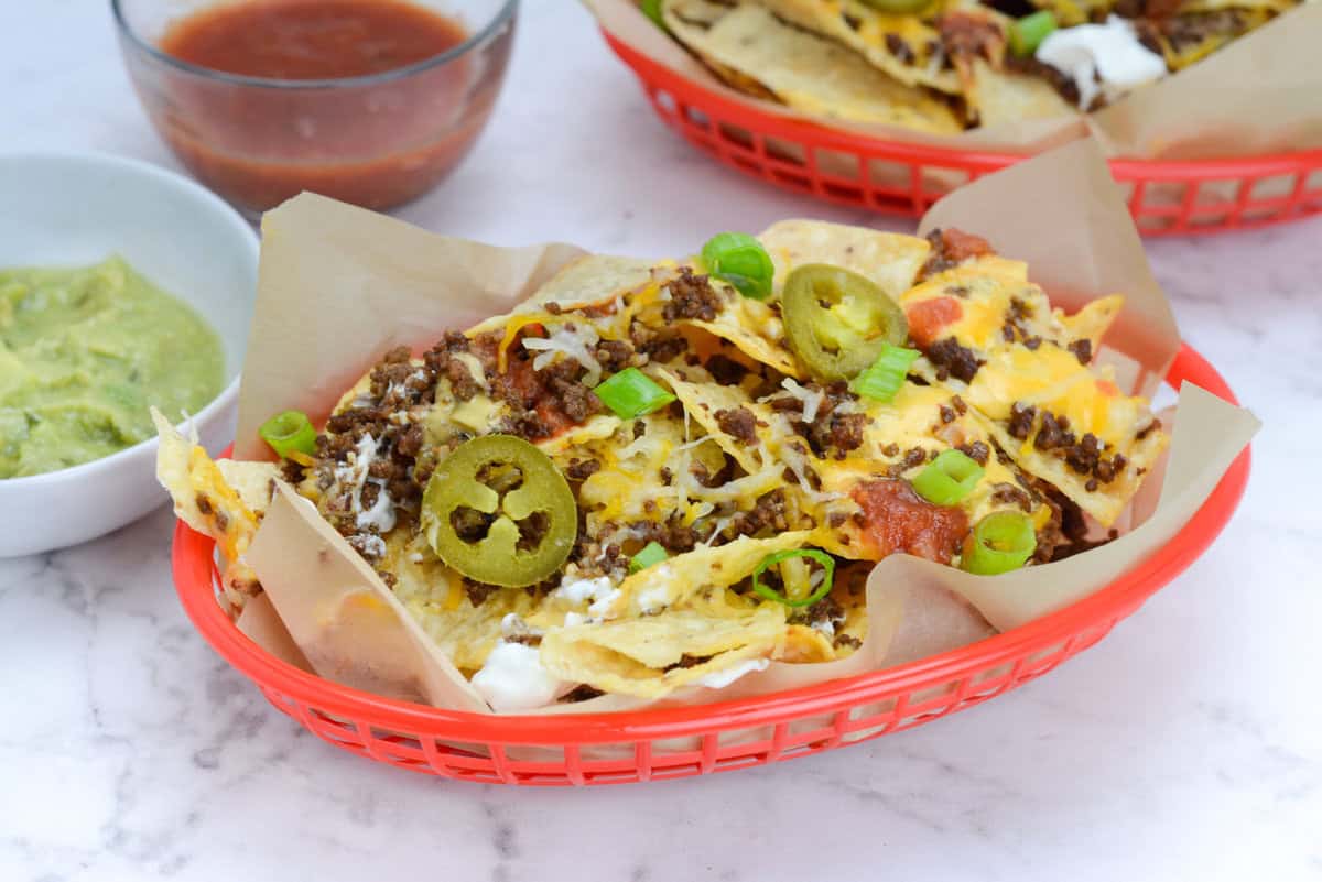 A red basket is lined with brown parchment paper. A serving of nachos is placed inside and topped with green onions and jalapenos. To the side is small bowls of guacamole and salsa and another nacho basket 