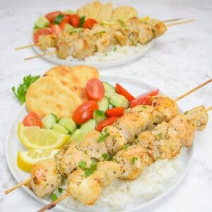 two chicken souvlaki skewers sit on a bed of rice. Beside them are two lemon slices, a cucumber and tomato salad and flat bread. Another filled plate is placed in the background.