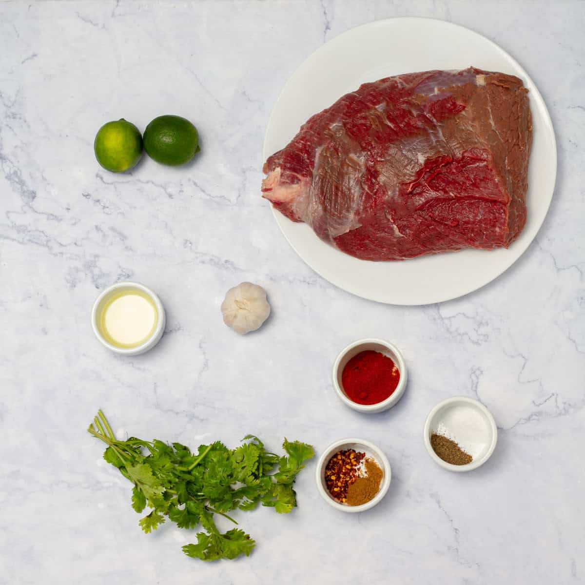 Carne asada ingredients of round steak, fresh cilantro, seasonings, limes, and oil in separate dishes. 