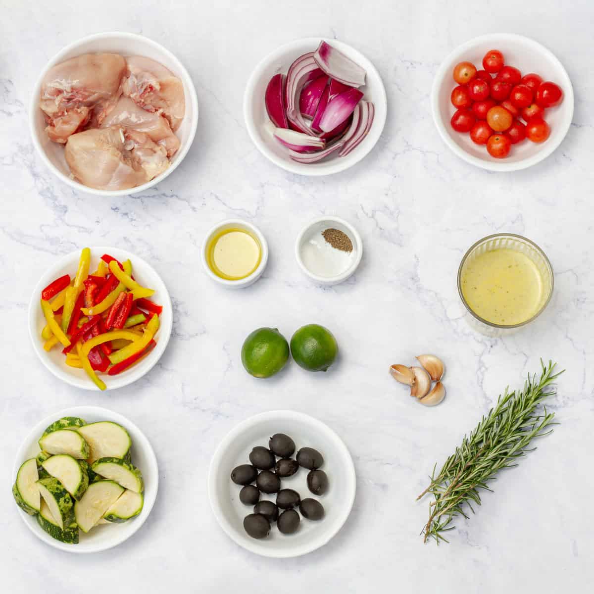 Greek Lemon Chicken ingredients of chicken breast, tomatoes, bell pepper, zucchini, olives, limes, garlic, sprigs, and and oil in separate dishes on a counter.
