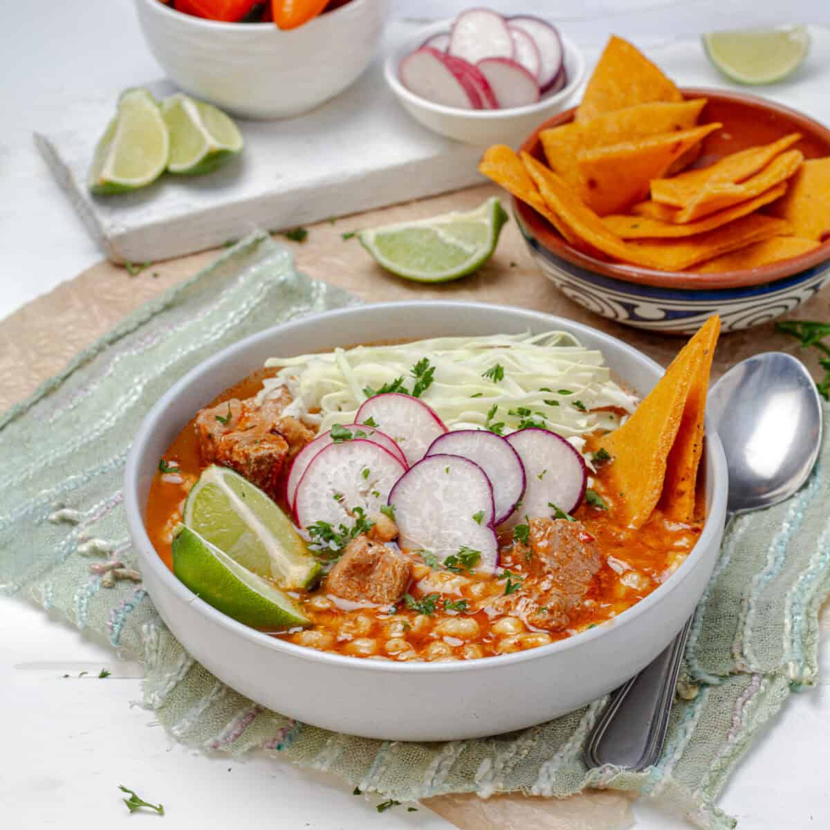 A bowl of light pozole made with hominy, topped with shredded cabbage, radish slices and lime, with tortilla chips behind it.