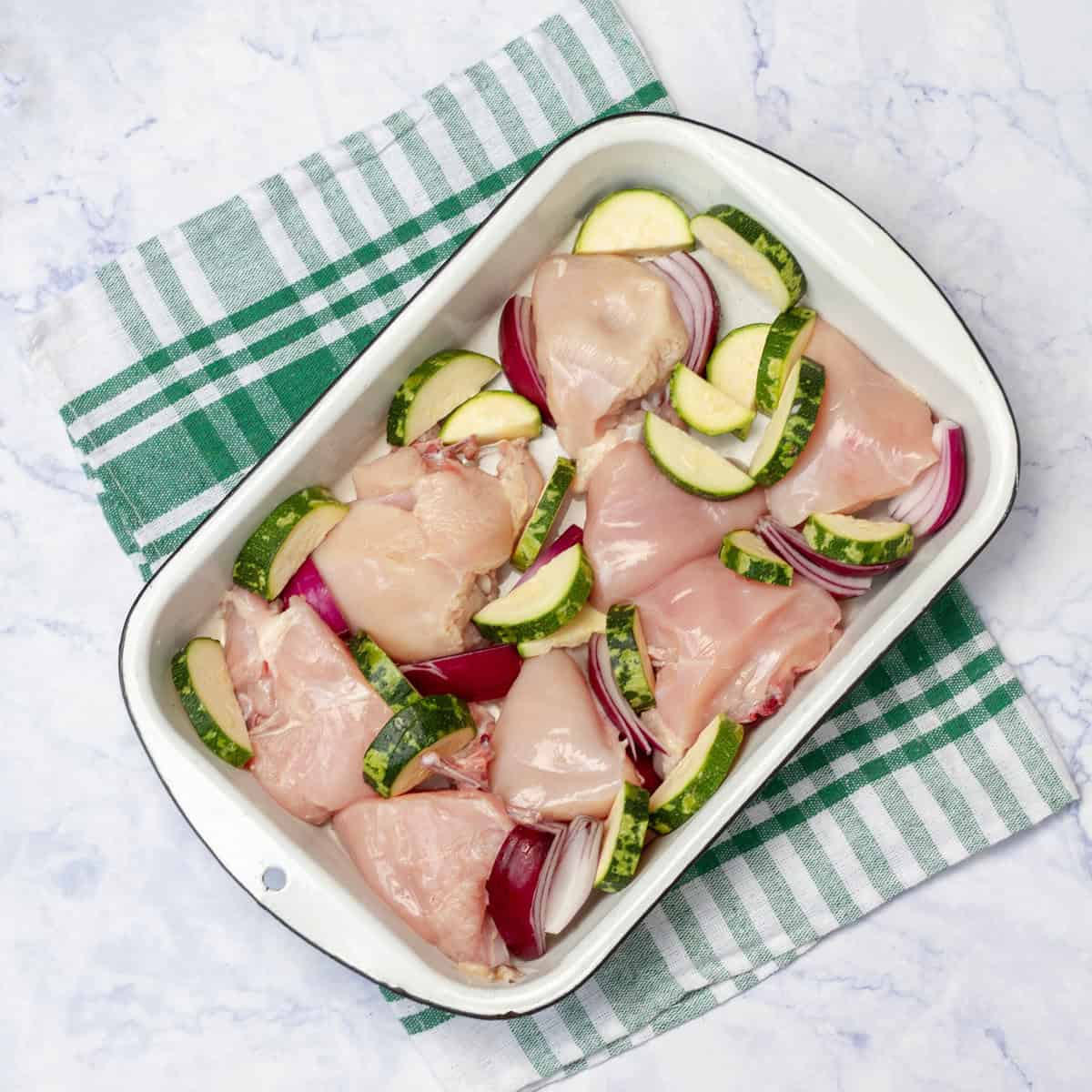 Raw chicken pieces sitting atop sliced zucchini and garlic in a baking dish. 
