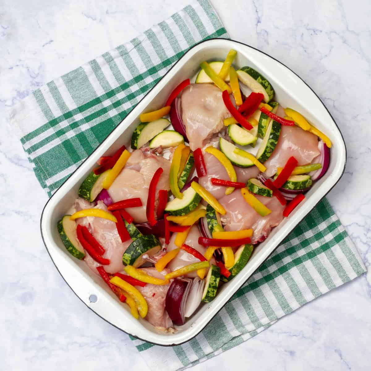Raw chicken pieces sitting atop sliced zucchini and garlic with sliced red bell peppers on top in a baking dish. 