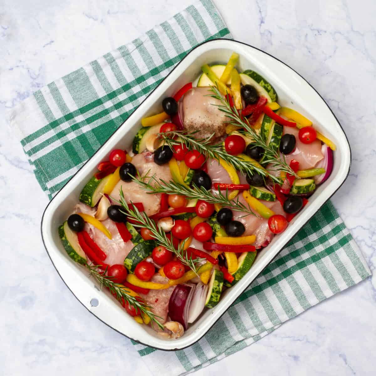Raw chicken pieces sitting atop sliced zucchini and garlic with sliced red bell peppers and black ilives on top in a baking dish. 