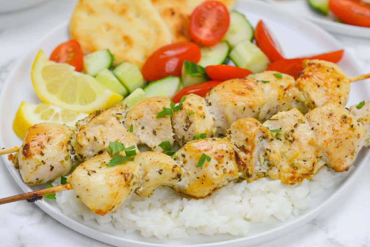 A close up of the cooked chicken souvlaki on a bed of rice. In the background is lemon wedges, a cucumber and tomato salad and bread. 