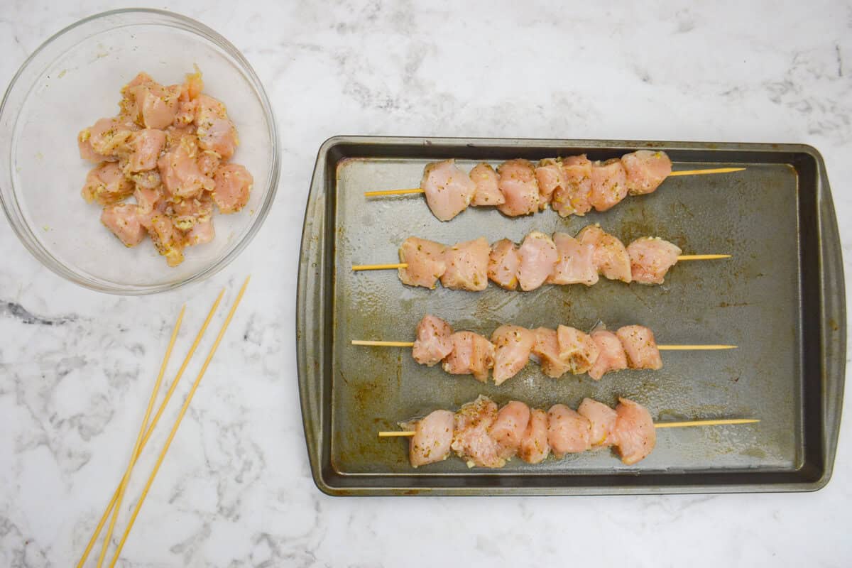 A medium sized clear bowl and skewers sit off to the side. In the center a baking sheet with four chicken skewers is shown 