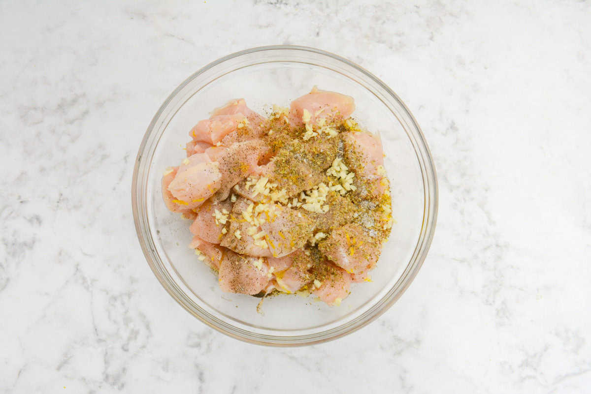 A large clear bowl is filled with diced chicken and topped with seasoning and minced garlic 