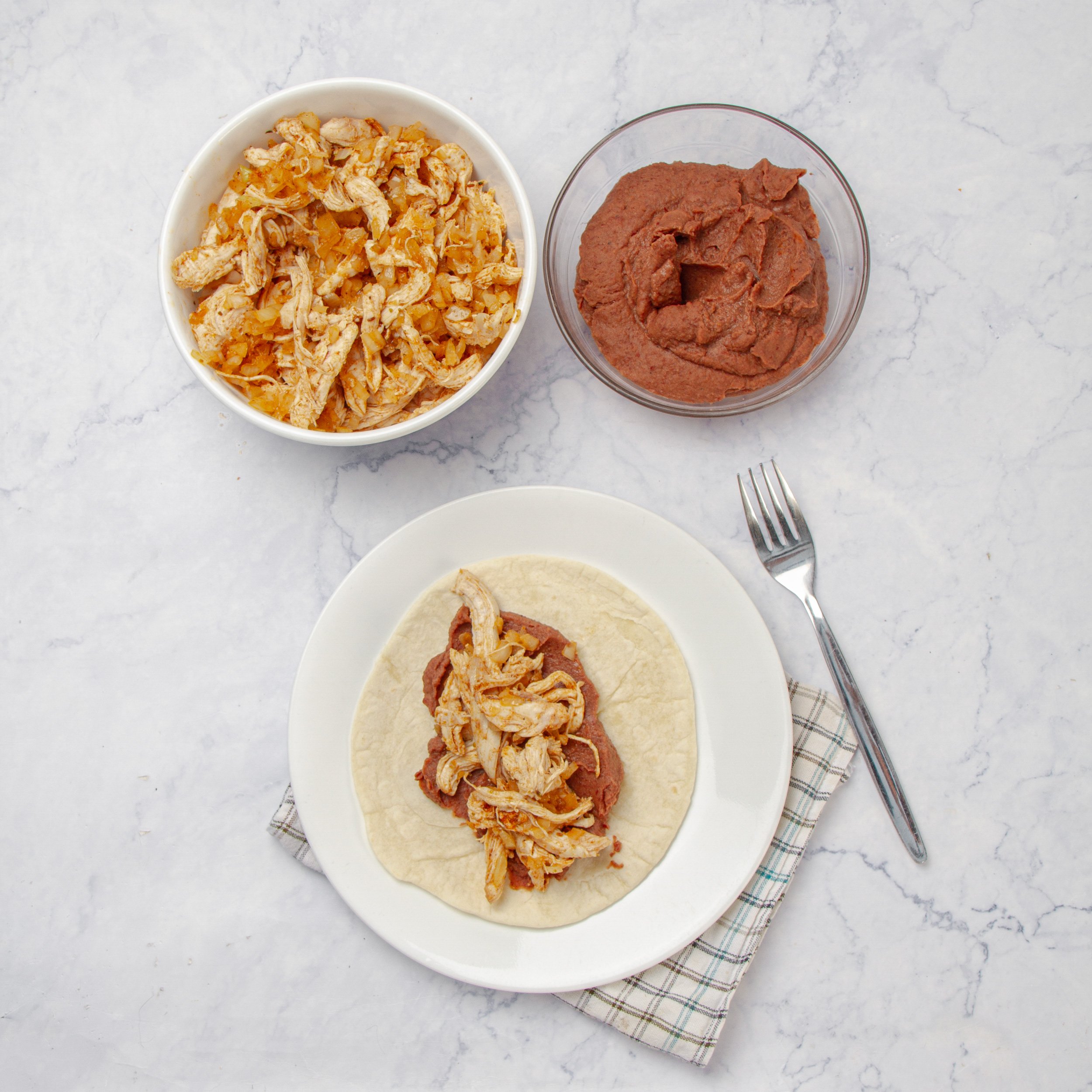 A tortilla being filled with refried beans and shredded chicken.