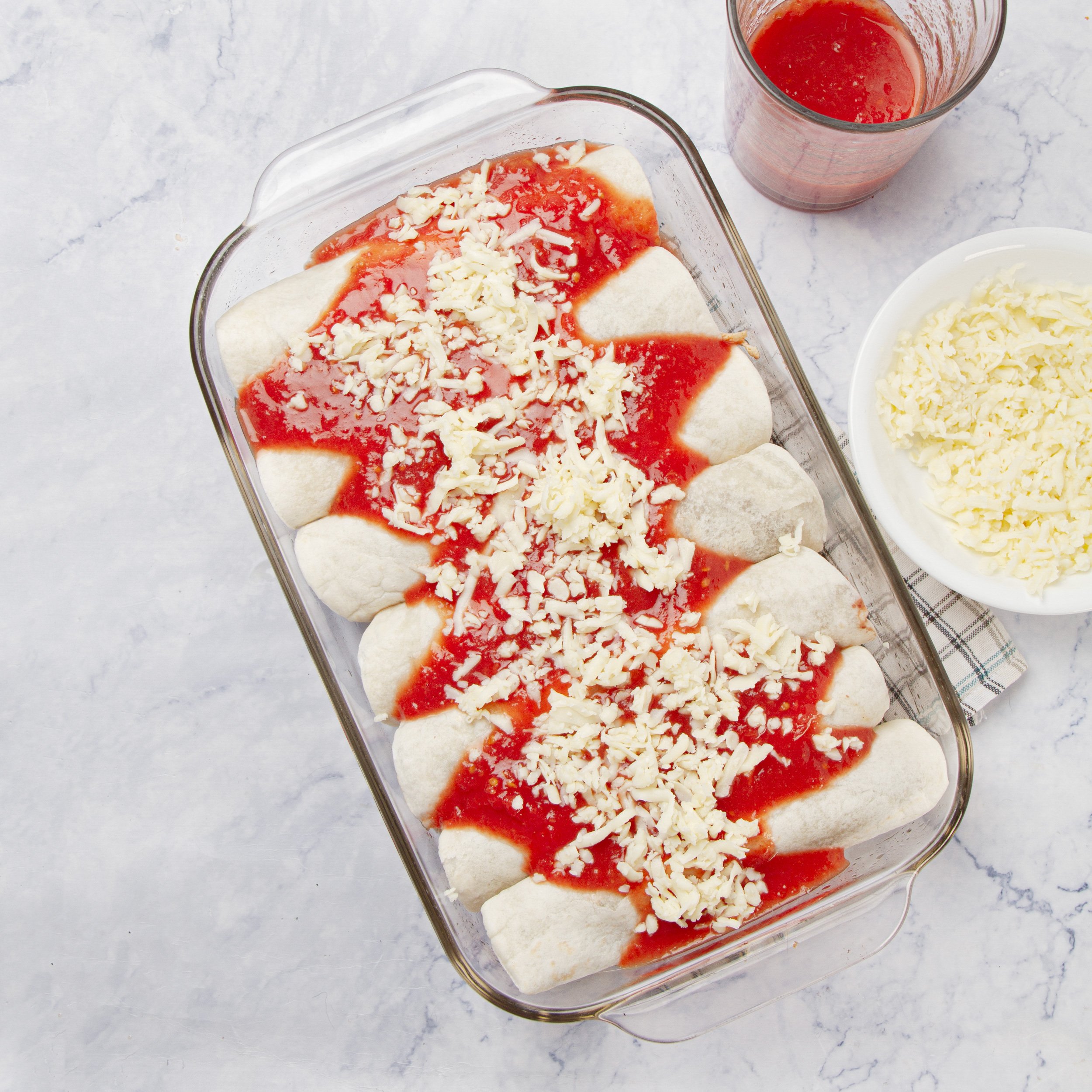 Filled and rolled tortillas fit snugly into a glass baking dish, smothered with sauce and shredded cheese.