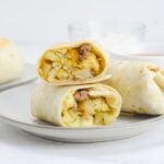 A cut breakfast burrito is on a plate. To the side, sits an uncut burrito and a bowl of salsa.