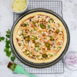 The cooked pizza is on a cooling rack and surrounded by small bowls with onions and cheese. Additional cilantro is placed to the side and a pastry brush with barbecue sauce is placed to the side.