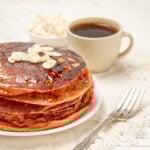 A stack of golden-orange Sweet Potato Pancakes on a plate with a fork and a cup of coffee on the side.