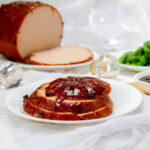 Slices of a beautiful traditional glazed ham on a plate with the whole ham in the background.