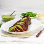 Juicy and tender London Broil sliced on a plate with green sauce drizzled on top.