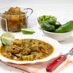 Thick and meaty Green Chili Stew in a bowl with whole peppers on the side.