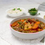 Hearty and comforting Milligan Stew in a bowl with a bowl of fresh herbs and rice on the side.