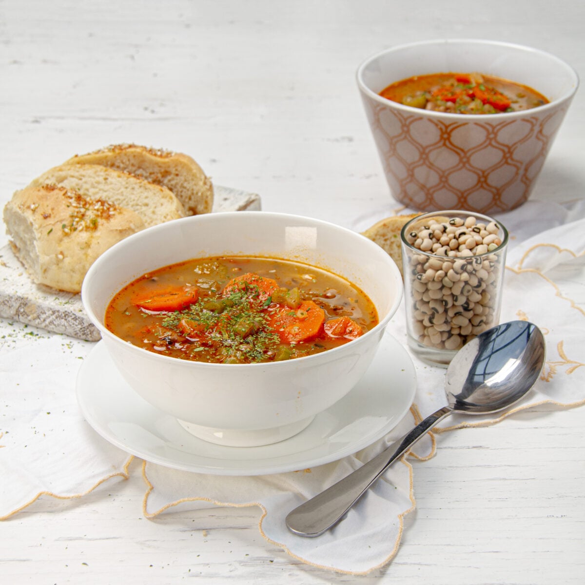 Black eyed pea soup in a white bowl with a jar of dried black-eyed peas, bread and a spoon beside it.