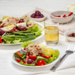 Fresh and flavorful Niçoise Salad on a plate with extra salad and a glass of juice on the side.