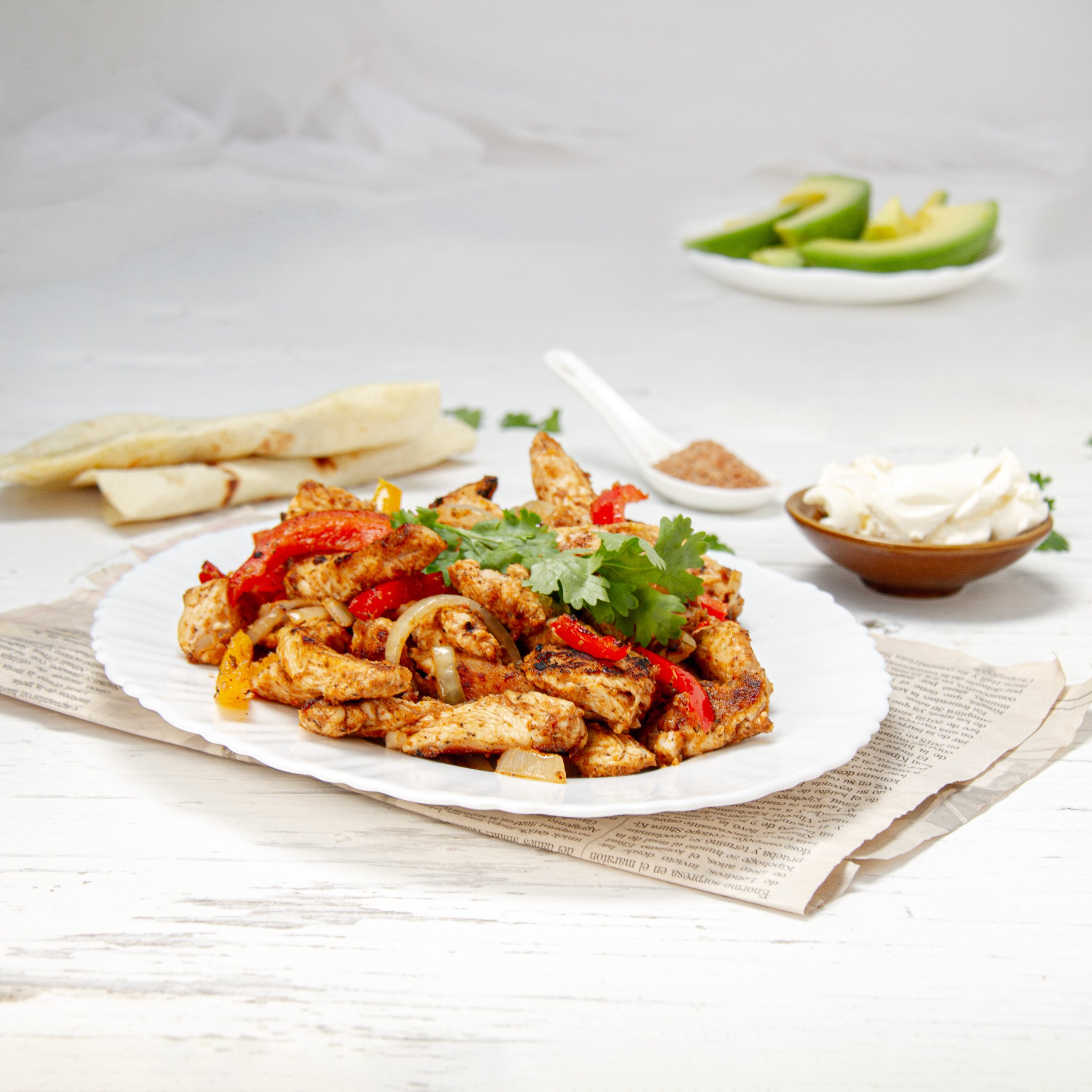 Sizzling healthy chicken fajitas on a plate with fresh cilantro garnish and avocado slices, tortillas, and sour cream on the side.