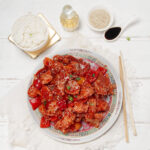 Healthy sweet and sour chicken bites on a plate with sesame seed garnish , rice, and chopsticks on the side.