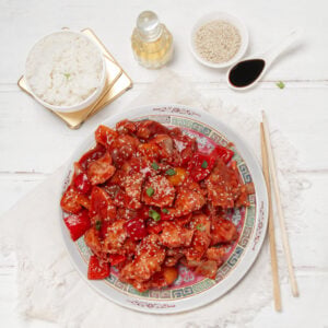 Healthy sweet and sour chicken bites on a plate with sesame seed garnish , rice, and chopsticks on the side.