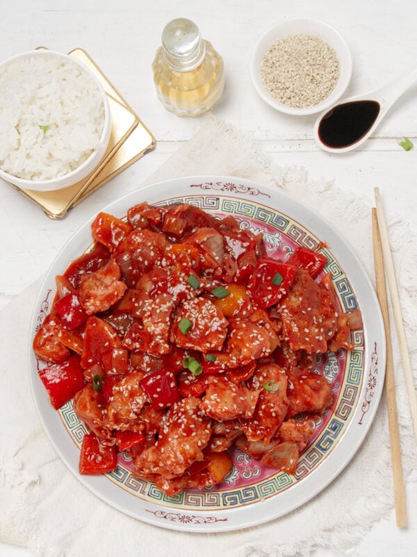 Healthy sweet and sour chicken bites on a plate with sesame seed garnish , rice, and chopsticks on the side.