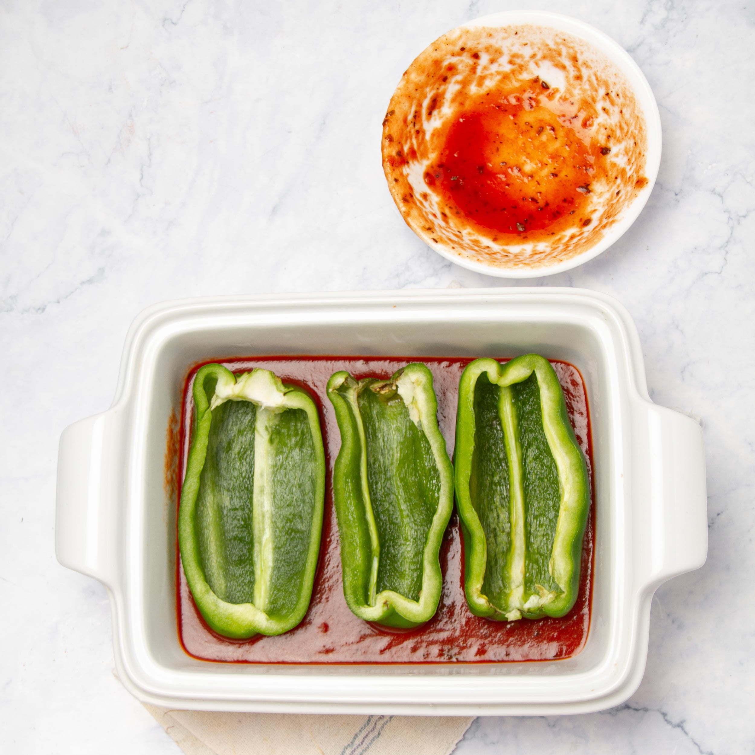 Three hollowed poblano halves placed cut side up on a layer of enchilada sauce in a white baking dish.