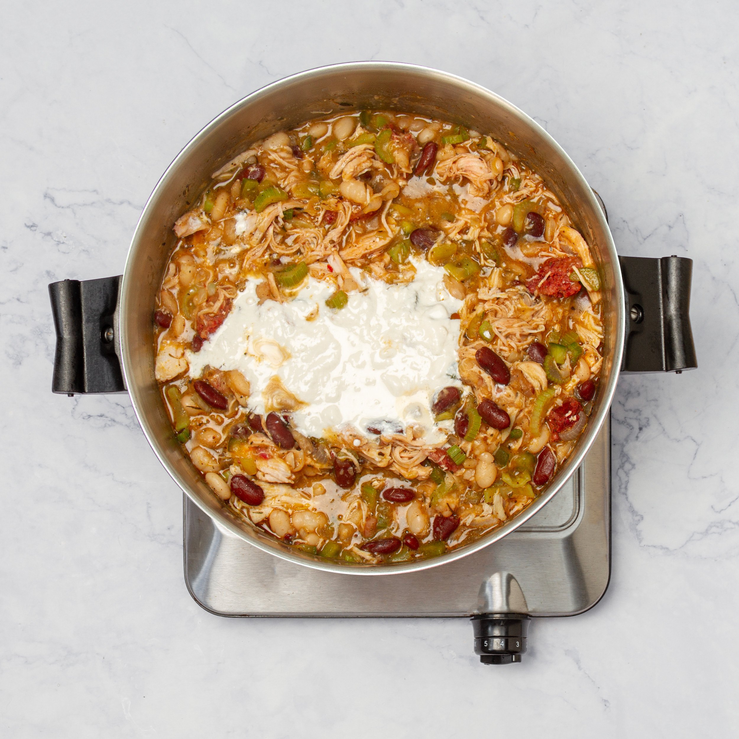 Chicken, white beans, and cheese added to the pot of chili on the stove.