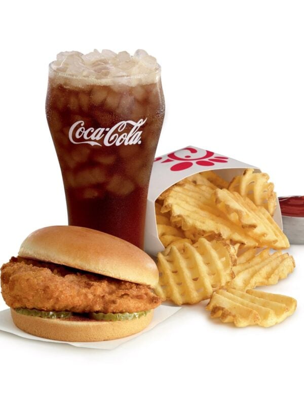 A Chick-fil-A Chicken Sandwich with pickles on a toasted bun, a carton of waffle fries with a cup of ketchup, and a glass of Coca-Cola over ice on a white background