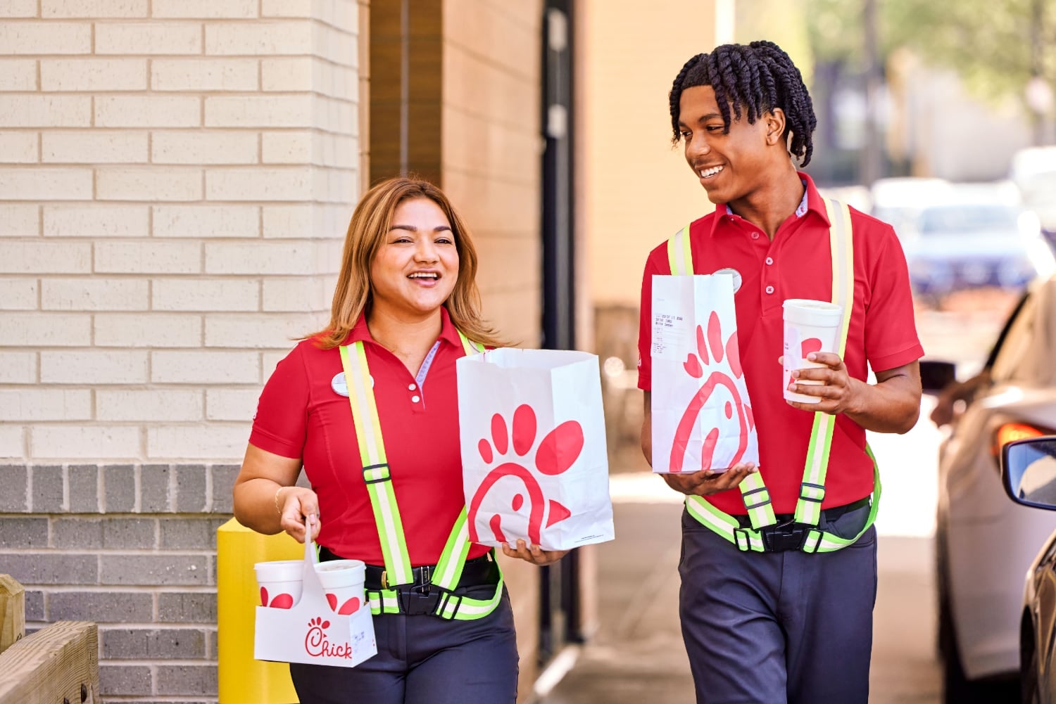Two Chick-fil-A team members in red polo shirts and safety vests carrying bags of food and drinks to cars in the drive-thru