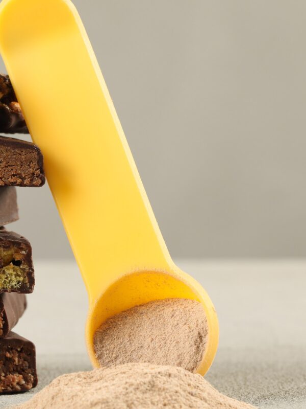 A stack of cut protein bars showing chocolate, nut and crisp layers beside a yellow scoop of protein powder on a light table