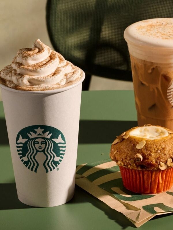 A Starbucks Pumpkin Spice Latte topped with whipped cream, an Iced Pecan Crunch Latte and a Pumpkin Cream Cheese Muffin on a green cafe table
