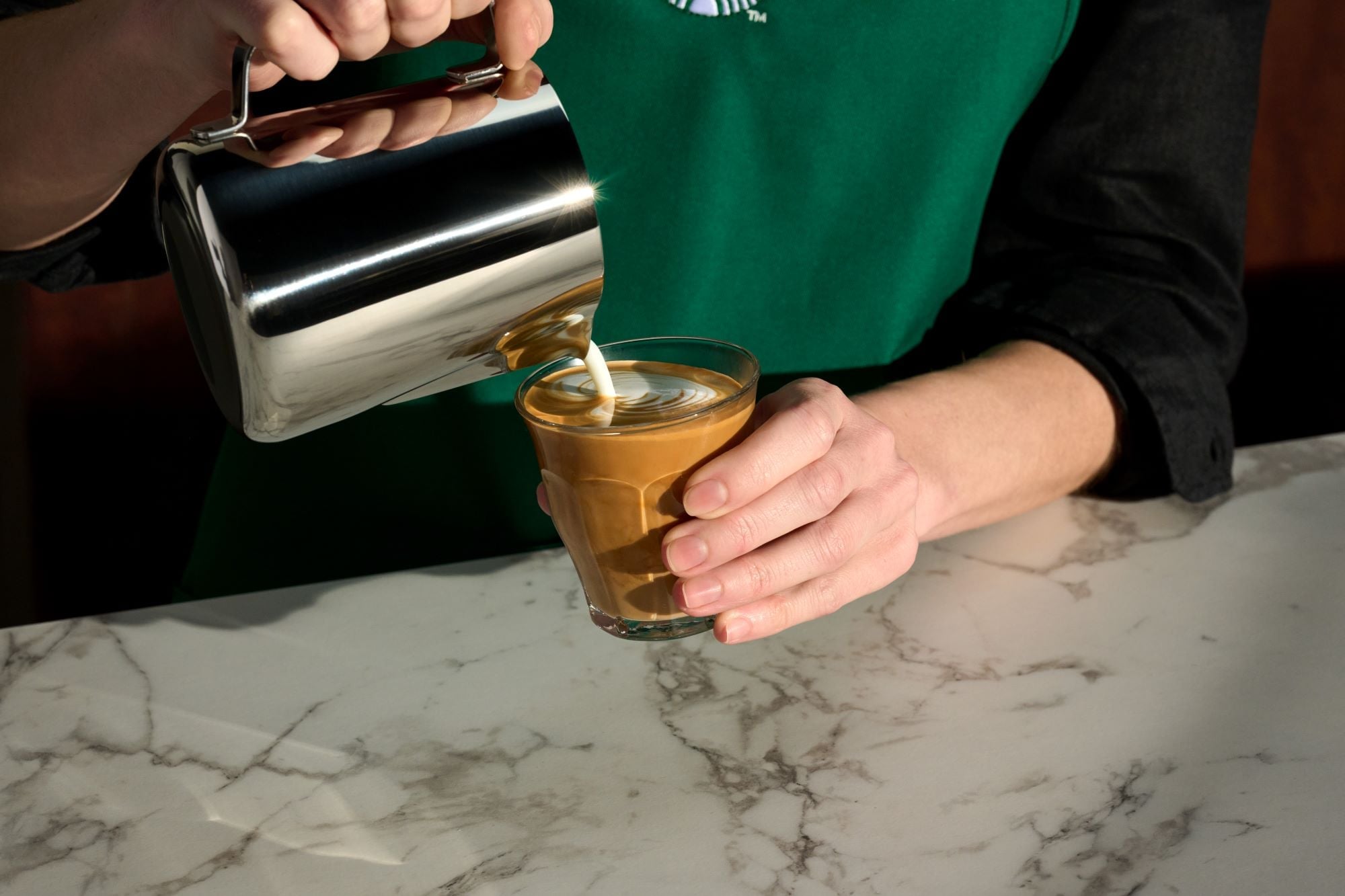 A Starbucks barista pouring steamed milk from a steel pitcher into a Pecan Cortado in a small glass