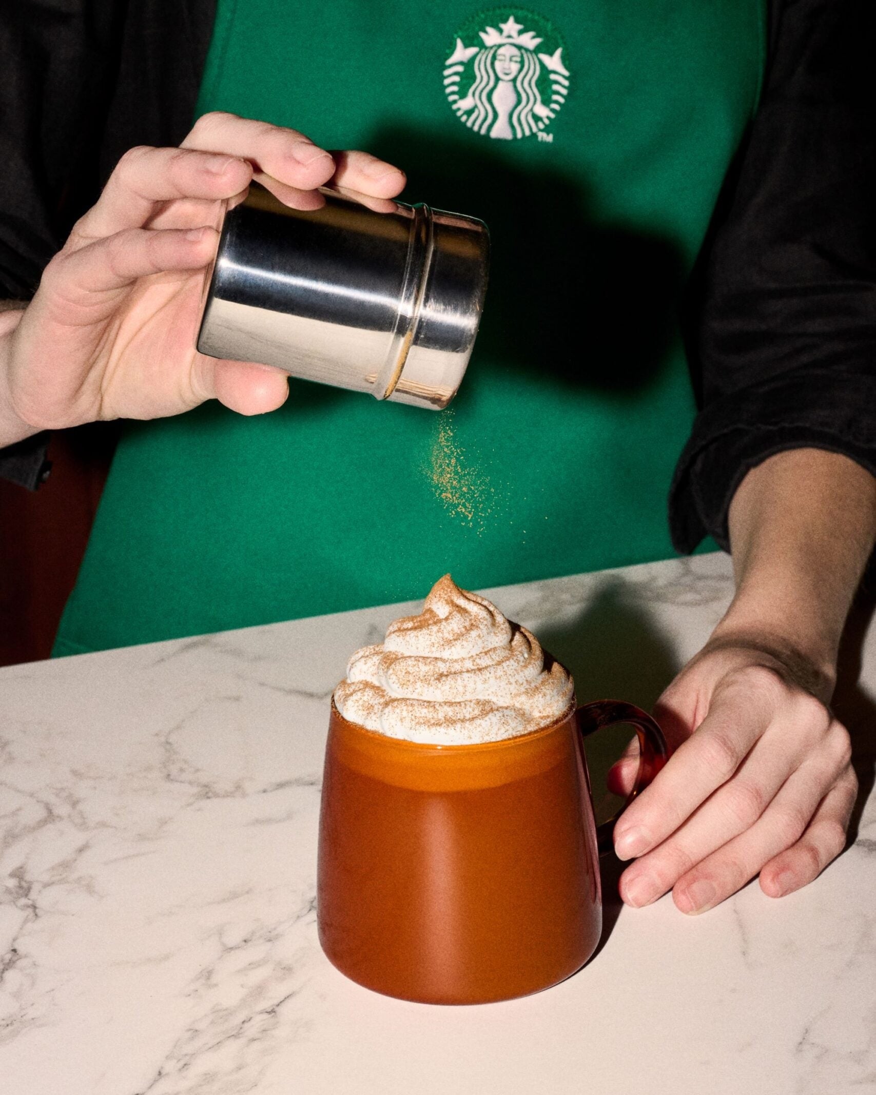 A Starbucks barista dusting pumpkin spice over the whipped cream on a Pumpkin Spice Latte in a clear glass mug